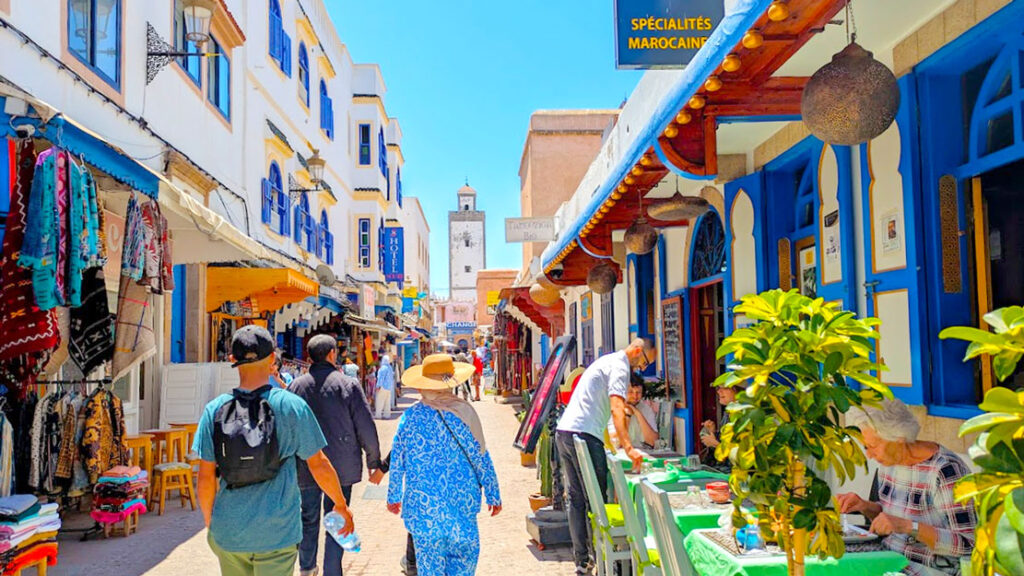 women and men walking away from the camera in the market in essaouria. Shops and restaurants line either side of the pedestrian filled street. The buildings are painted white with blue accents around the doors and windows. In the distance, above the stores and people walking is a mosque tower. 

More importantly, the woman is fully covered, wearing a blue patterned long sleeve pant outfit and a wide brim straw hat. The man beside her is in a short sleeve shirt, you can't see what pants he's wearing.