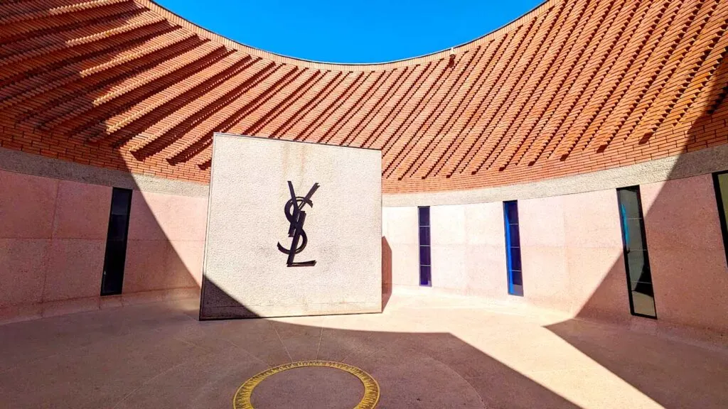 the entrance to the Yves Saint laurent Museum. A rounded courtuard in terracota colours. There are tall rectangle windows evenly spaced out. But in the centre is a wall with the Yves Saint Laurent Logo (Y S and L all intertwined