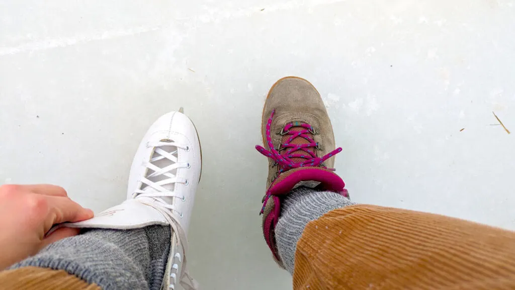 a top down view of a woman's legs (wearing corduroy pants) very warm pair of black socks, and her feet. On the left foot is a pair of figure skates, you can see her hand is still putting it on. The right food still has columbia newton hiking boots.