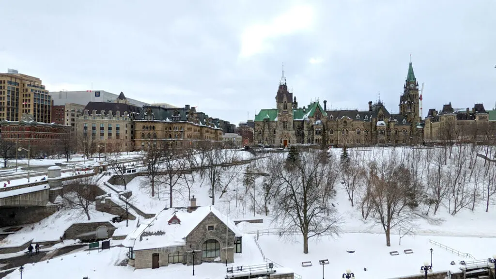 ottawa view of parliament and the rideau canal in front of it in winter. The sky is overcast, but the light is trying to peak through the clouds. There's a nice layer of snow covering the grass, trees, and buildings, and the canal in the middle - not ready for skating.