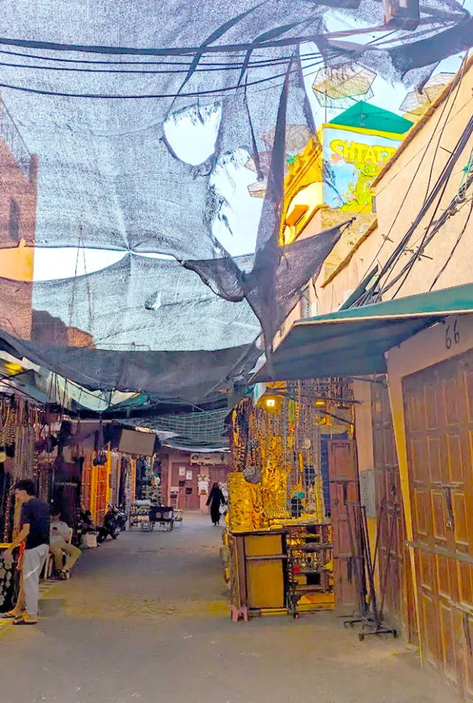 A less busy street in the Marrakech medina. There's a thin black cloth roof, that you can still see the sky through because it's ripped. The right side of the street, are all closed wooden doors. On the left, there are some vendors selling jewellery. In the distance you can see some tourists walking