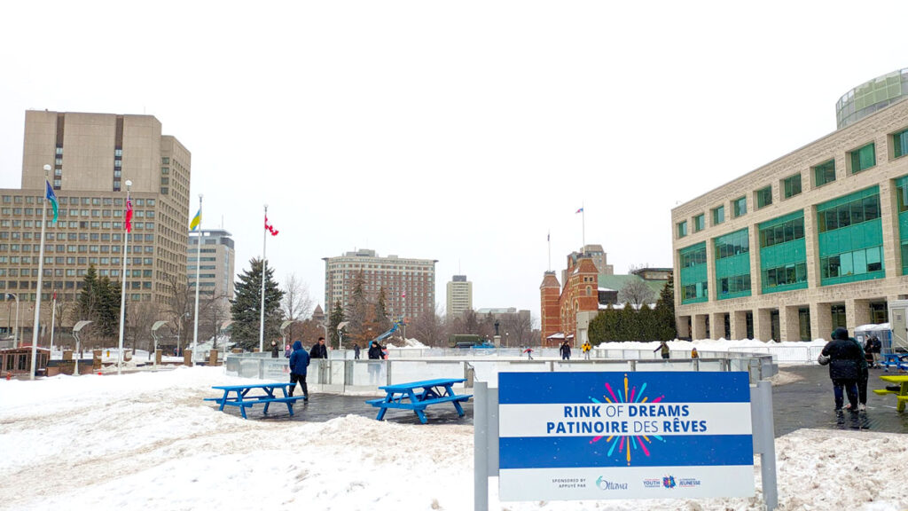 a sign in the foreground say: the rink of dreams. Behind it there's snow, with a cleared path to blue picnic tables. Then there's a large rink, with some people skating on it. there are buildings surrounding the rink, this is downtown Ottawa afterall