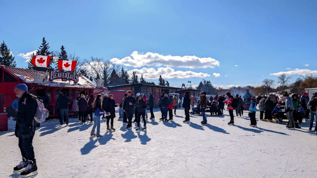 the lineup for Beavertails on the rideau Canal, with two large and proud canadian flags waving in the wind above it. It's a beautiful sunny day, with barely a cloud in the sky! People are in their skates in line, and enjoying the food. 