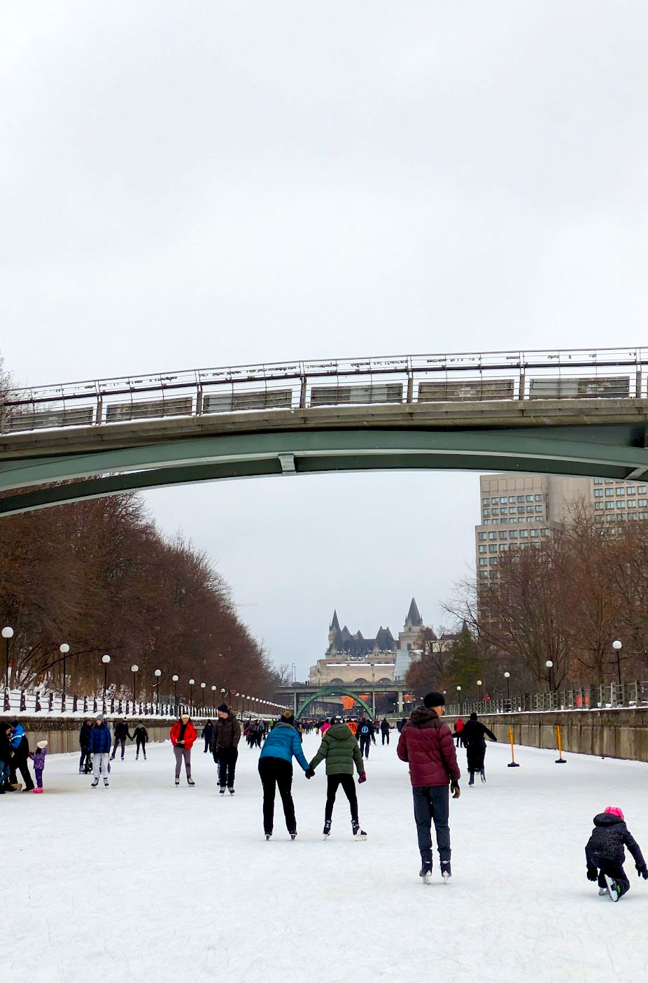 a view of people skating in the middle of the rideau canal in winter. A couple is holding hands directly under the bridge ahead. Concrete sides and lampposts line either side of the canal , and bare trees. In the distance is the castle of the chateau laurier. This is the best view while skating on the rideau canal. The sky is white as it's snowing.