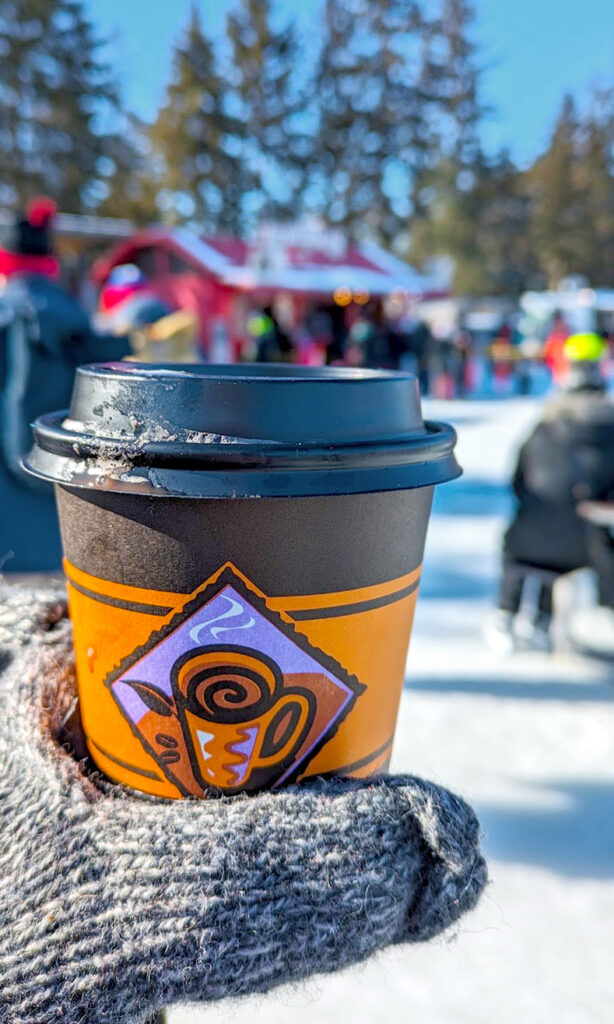 someone wearing knit gloves is holding up a to go coffee cup in the foreground. Out of focus on the background is a winter scene of people on ice with skates, food stalls, and evergreen trees