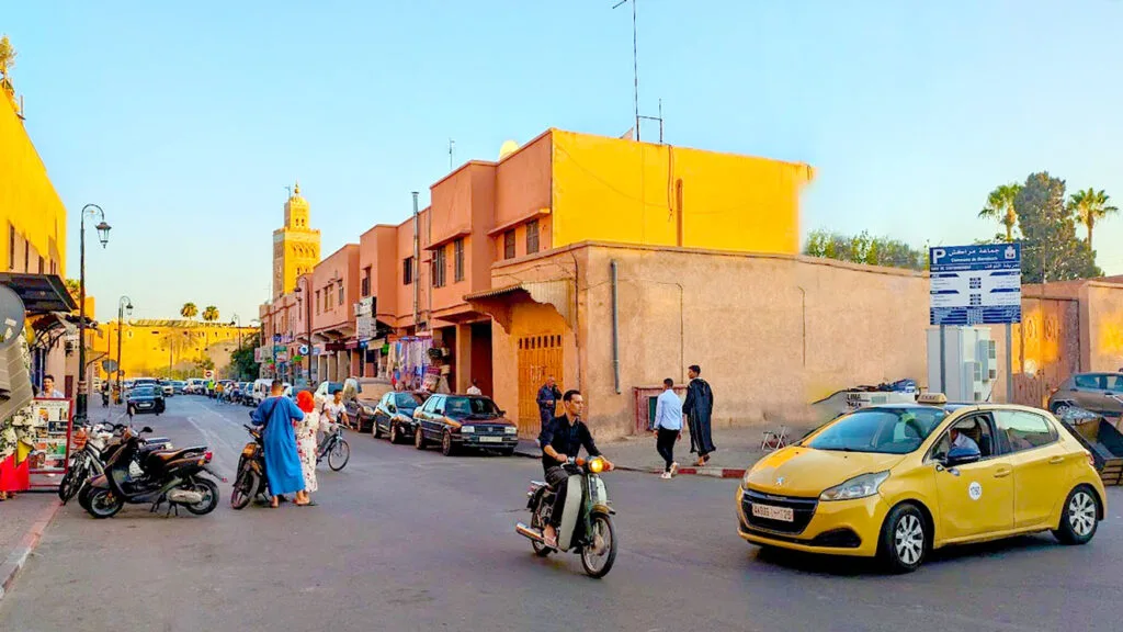 a busy intersection in old marrakech just outside the market. People are trying to cross the street, they're wearing bright royal blue full length dress, and bright white. A motorcycle is about to go into a yellow taxi in the middle of the intersection. It's golden hour, the setting sun is beautifully lighting up the orange facade of the buildings. Poking out above the buildings is the famous koutoubia mosque.