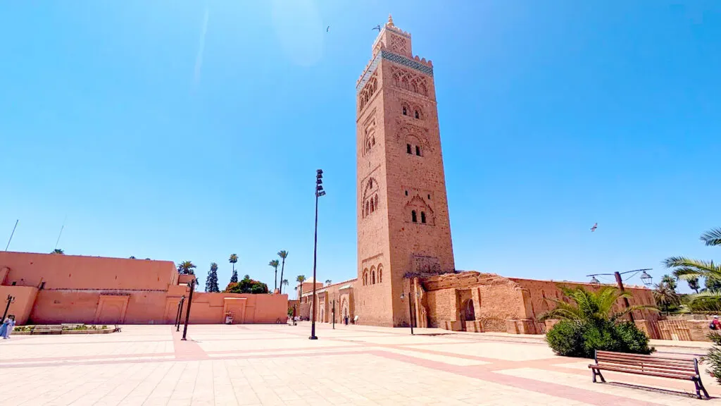 a large open courtyard in front of a tall tower, the koutoubia mosque in marrakech. There's beautiful vibrant colours, the ground stone is a pinkish hue, the facade of the koutoubia is a pinkish orange, and the sky is a crystal clear blue without one cloud