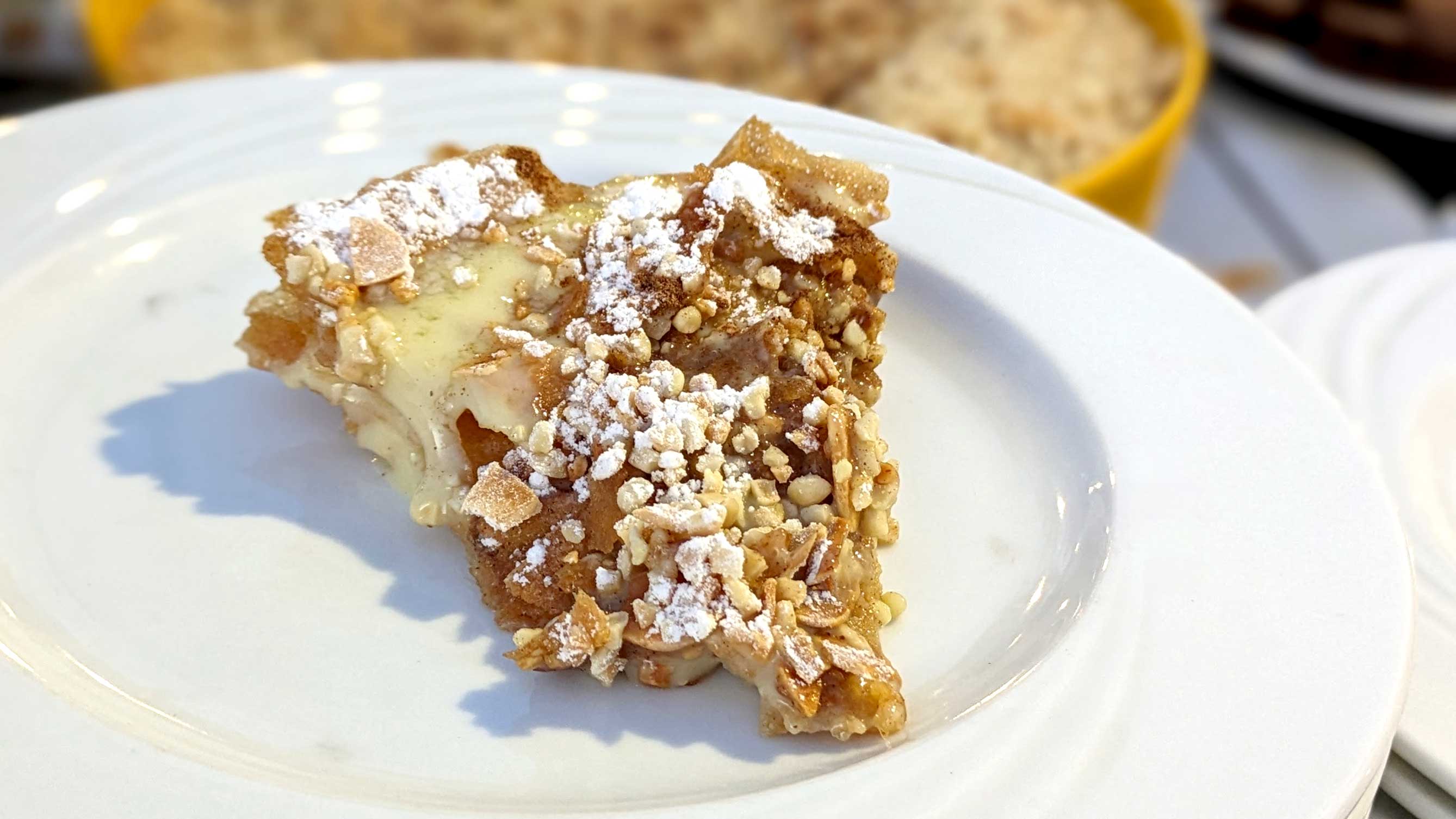 a white plate with a piece of Ktefa (milk pastilla) in the middle. It looks like a piece of pie but it's thin crispy pastry layers with a cream filling. You can see crispy fried almond pieces on top with powdered sugar