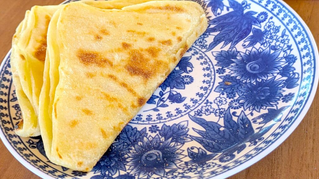 Two msemen sit on a blue and white floral patterned plate. They are folded over to create triangles. If unfolded they would be a square. The dough is nicely cooked, a soft yellow, with a few brown spots throughout the bottom 