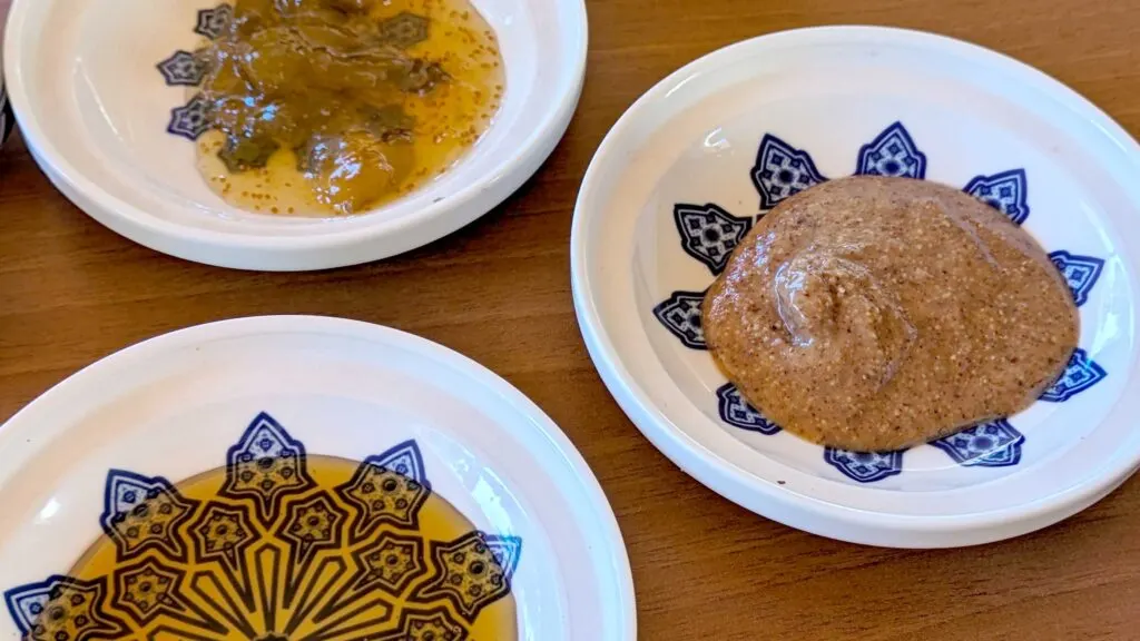 Three blue and white patterned plates on a tables. The farthest plate is an orange yellow jelly with seeds in it. The second below it has the bottom half of the plate cut off, it looks like honey is on the plate. The last plate is amlou, it looks like natural almond butter, the colour of nut butter with little tiny bits of almond in it. It also has a shine to it, probably from the oils