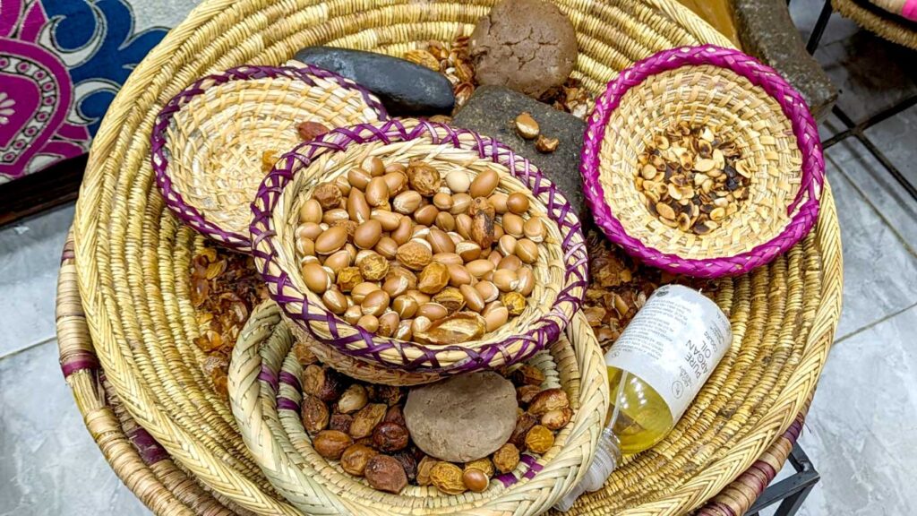 a large straw bawl is in the middle of the frame. It's filled with smaller straw bowls with colorful rims, from purple to pink and yellow. Each one is filled with a kernal from an argan tree at different stages. Thre are whole perfect looking shiny kernels, wrinkly shells, and then roasted pits. There's also a bottle of argan oil in the large straw bowl