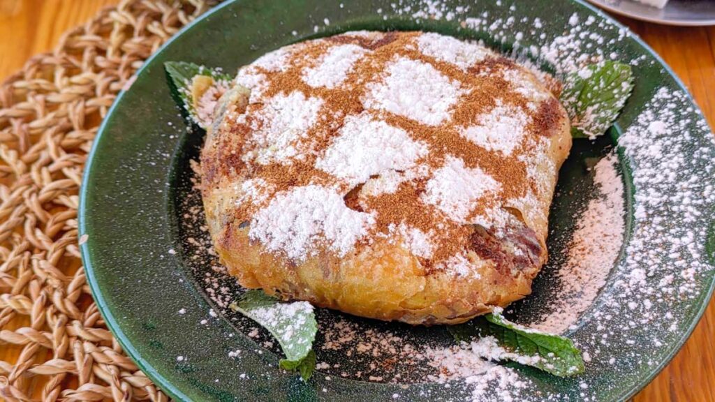 A b'stilla on a plate. a circular stuffed filo pastry , there;s an argyle pattern made with powdered sugar and cinnamon. Below the pastry are fresh mint leaves.