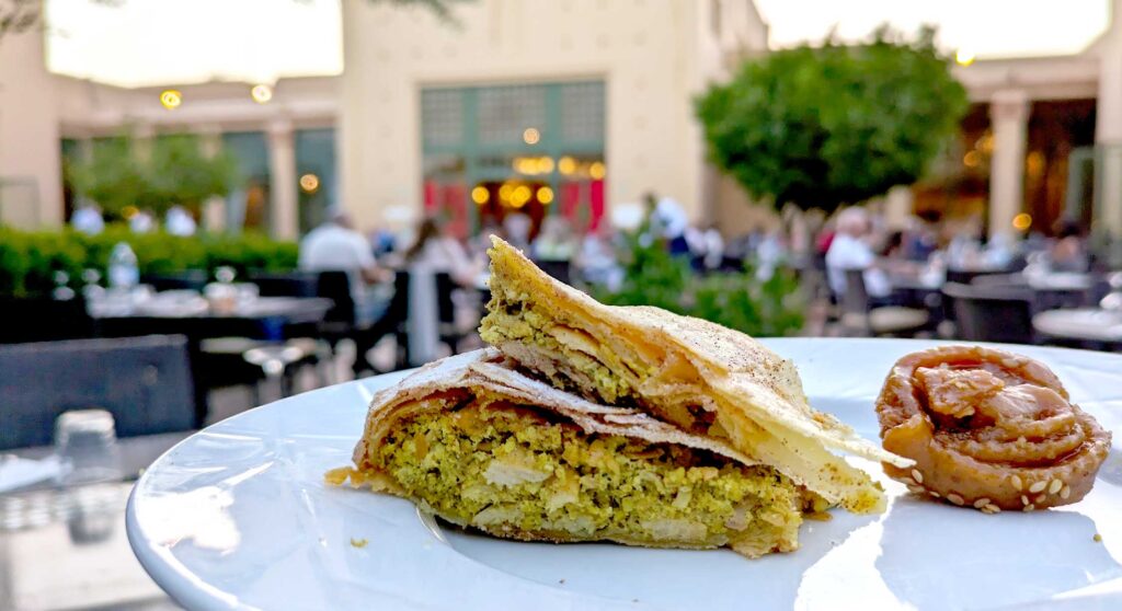 a plate filled with two pieces of chicken pastilla and a chebakia. The pastilla looks like a covered pie with chicking filling. The chebakia is a rose shape dough ball glazed with honey and sesame seeds. In the background is a pastel moroccan building with trees and tables.