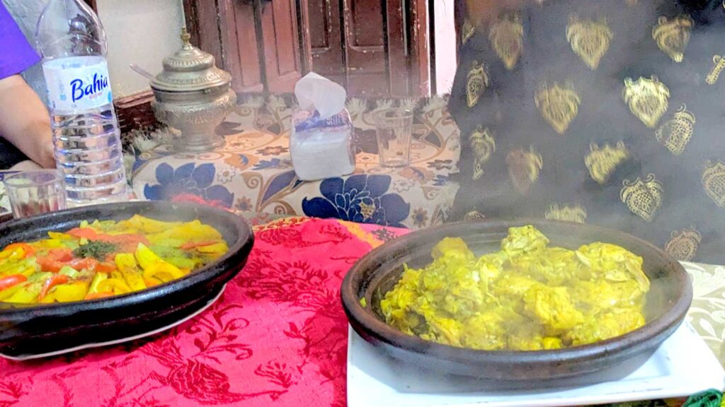 two tagine plates on a red table cloth in a local home in Morocco. You can tell the lids you removed only seconds before because there is so much steam coming off of the dishes. In the dish on the left is a beautiful patterned vegetables, all different colours from red, yellow, orange and green. The tagine on the right has chicken with preserved lemon. It has a rich yellow colour, but the meat is so tender you can see that it's already fallen off the bones.