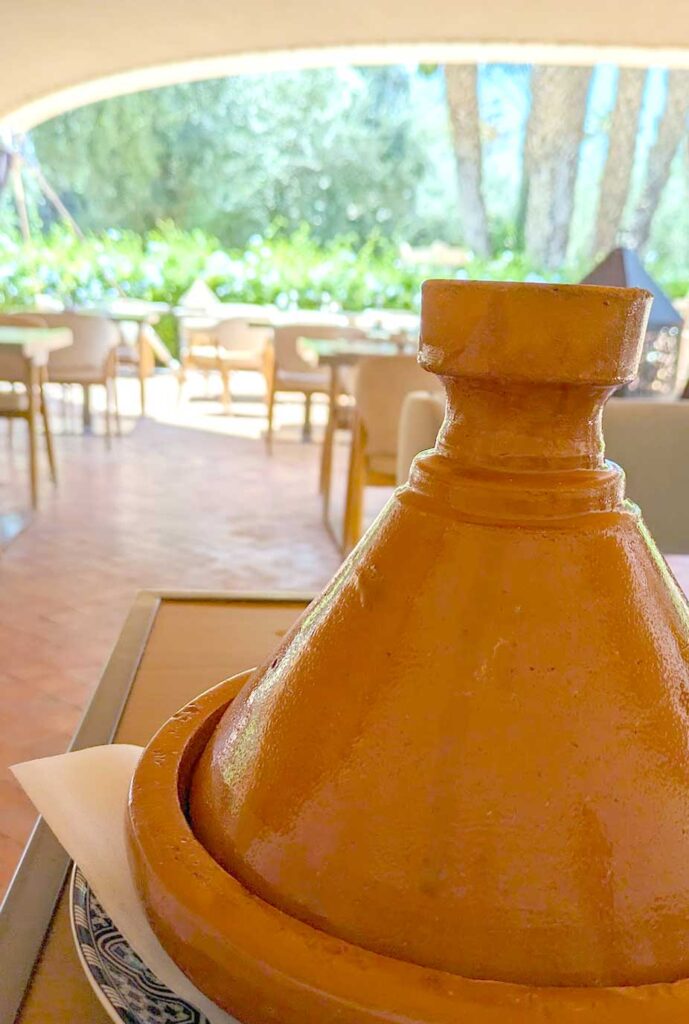 a vertical image. The background is out of focus, it's and outdoor restaurant, the sun is starting to peak in where it isn't covered. On the table in front of the camera sits a large terra cotta tagine. It's a burnt orange colour, but very basic. It's still covered so you don't know what's inside. 