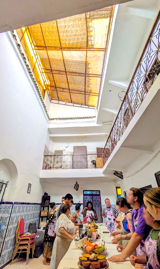 a vertical image showing the inside of a moroccan house with a cooking class taking place. There's no solid roof, but bamboo sheets with small holes to let light in. A long table is set up on the first floor in a courtyard of the house. People are standing around, all wearing the same pink apron. The chefts stand on the left side of the table, giving instructions. Ingredients were already laid out in front, including spice bowls and a bowl of raw vegetables. Cutting boards are also placed in front of each particiapnt