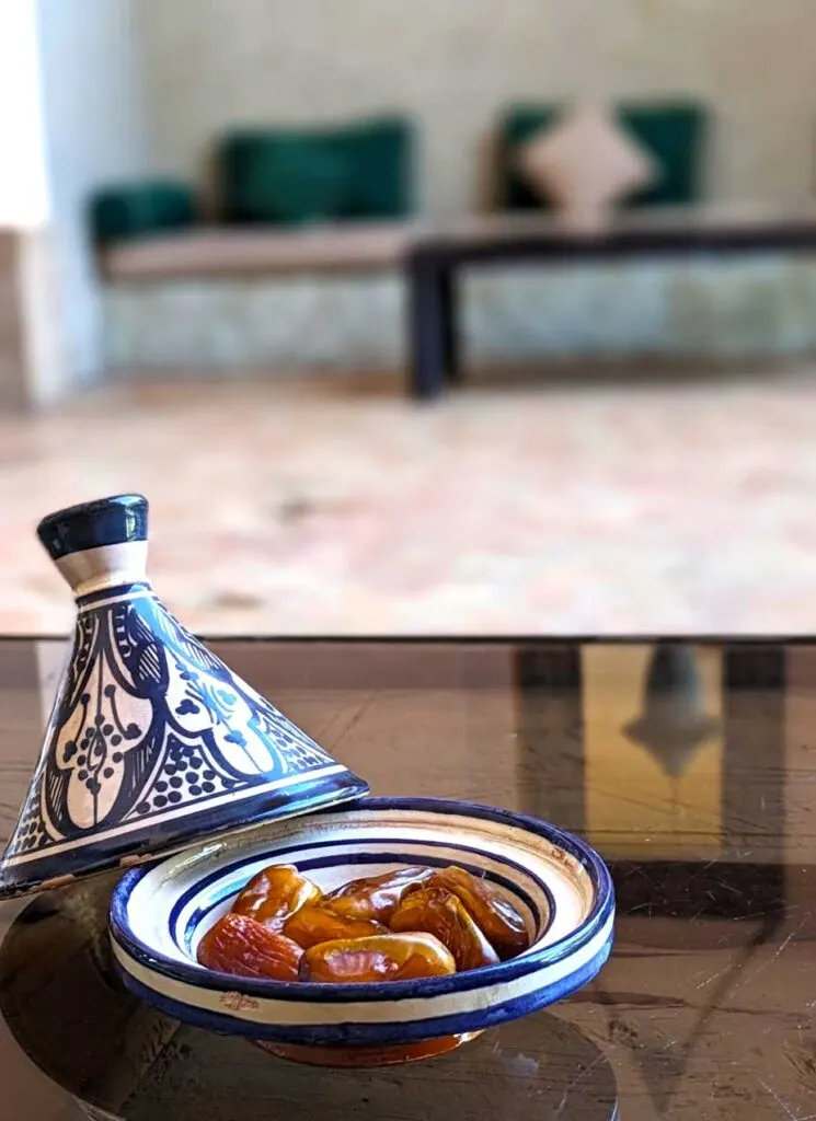 a blue and white mini tagine filled with dates. The cover of the tagine is resting on the bowl, on a bit of an angle. The tagine is on a glass top coffee table, so you can see the reflection of the room on the table There's a moroccan lantern and a doorway. Across the table is out of focus, a couch with evergreen pillows and the same coffee table