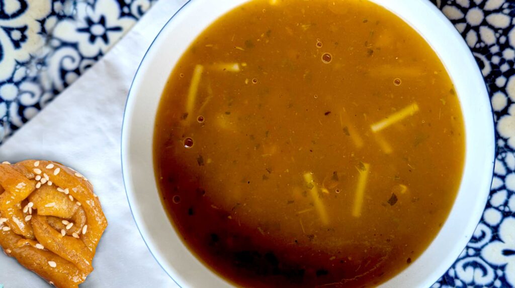 an overhead horizontal photo of a bowl of tomato soup with noodles. Next to the bowl is a rose shape dough ball with sesame seeds on the outside and a glazed honey look. Underneath the bowl is a napkin and a blue and white patterned table