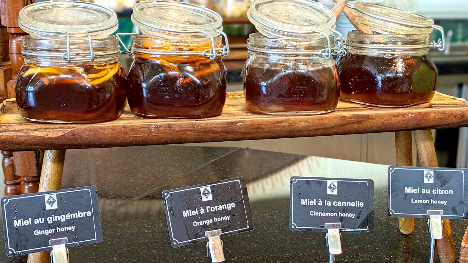 four jars of honey sit on a shelf, each one is a deep rich dark golden colour. 

Below, are table cards with what each honey are. 

The first jar has yellow strips in it, it's ginger honey. The second has more orangy strips, orange honey. The last two just look like regular honey but the labels says the third is cinnamon honey and the last one is lemon honey. 