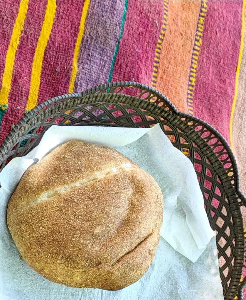 a loaf of moroccan bread khobz sit in a brown bread basket lined with napkins. The basket sits on a moroccan blanket, with yellow, red, purple, and orange vertical stripes