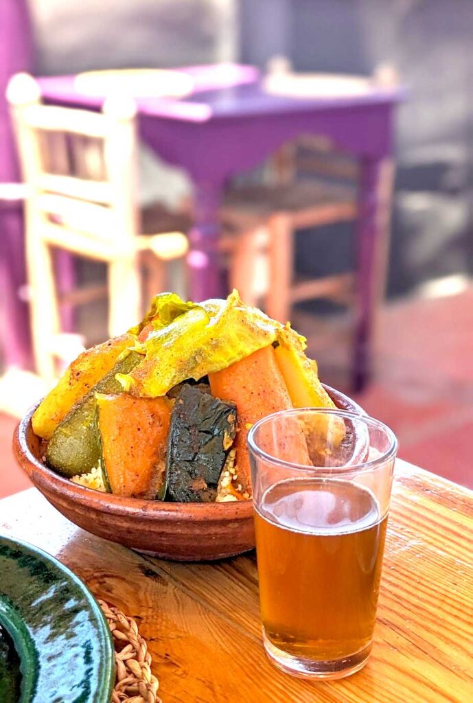 vertical image. a table at a restaurant. A purple table is blurred out in the background with two chairs.

The foreground has a couscous meal and a glass filled with hot mint tea. The couscous is mostly covered by a pyramid of vegetables, from squash to zucchini and a piece of lettuce on top.  