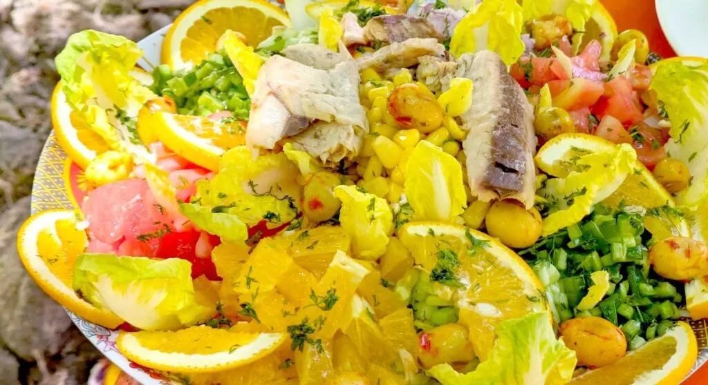 close up of a freshly made salad. Lining the edges are orange slices and pieces of lettuce. There are piles of veggies like green peppers, greenonions, cucumbers, and tomatoes around the side, in the middle is a pile of sweet corn, and on top is cooked mackerel