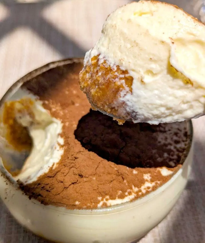 tiramisu-a-amlou-moroccan-desserts-to-try. A glass bowl with a white mousse inside and cocoa powder on top. There's a spoon size hole in the bowl, and a spoon is on top of the bowl with the contents. You can see the cream mousse and amlou underneath