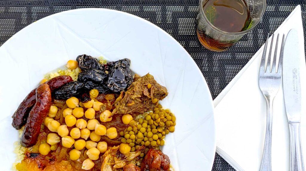 an overhead view of a white plate filled with traditional moroccan foods to try. Sitting on a bed of couscou, there are two short, narrow, and deep red merguez saausages, a pile of prunes, beef, green lentils, som chicken and dried apricots hidden underneath some chickpeas. It's on a black patterned table settings, there's a fork and knife placed on a folded napkin next to the plate and near the top of the photo is a glass of hot mint tea