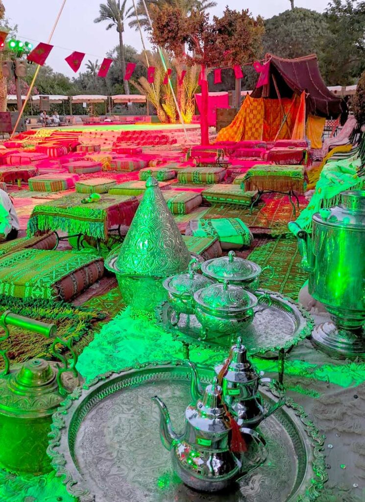 two moroccan tea pots sit on a large silver tray. Behind the teapots is the set of an arabian night. There are pillow on the floor for people to sit and small tables. There are red and green lights to set the scene and there's a stage in the distance. Palm trees hang above in the dusk sky. 