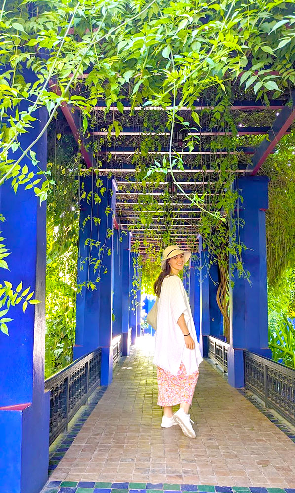 a woman wearing a white shawl and maxi pink and orange dress with a sun hat stands looking back smiling towards the camera. She stands under a covered pergola that's painted indigo blue and covered in green leaves