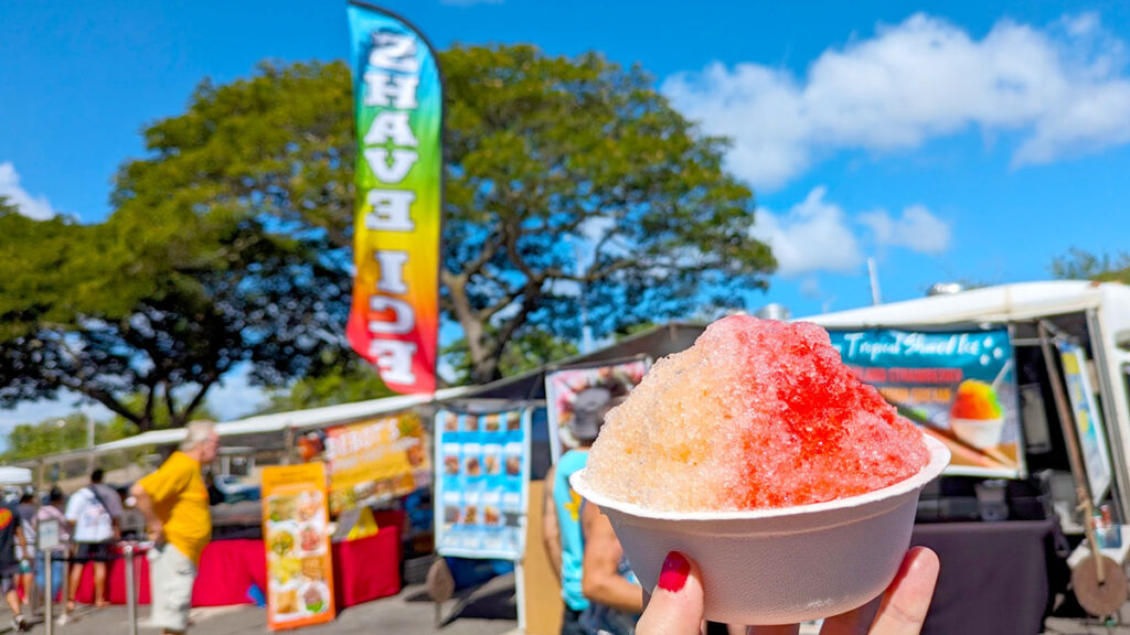 a hand holds up a bowl filled with crushed ice (or shave ice). It's in a domed shape and split down the middle the left side is orange and the right is red. In the background, slightly blurred is a bright shave ice sign blowing in the wind. The background colours of the sign are the colours of the rainbow.