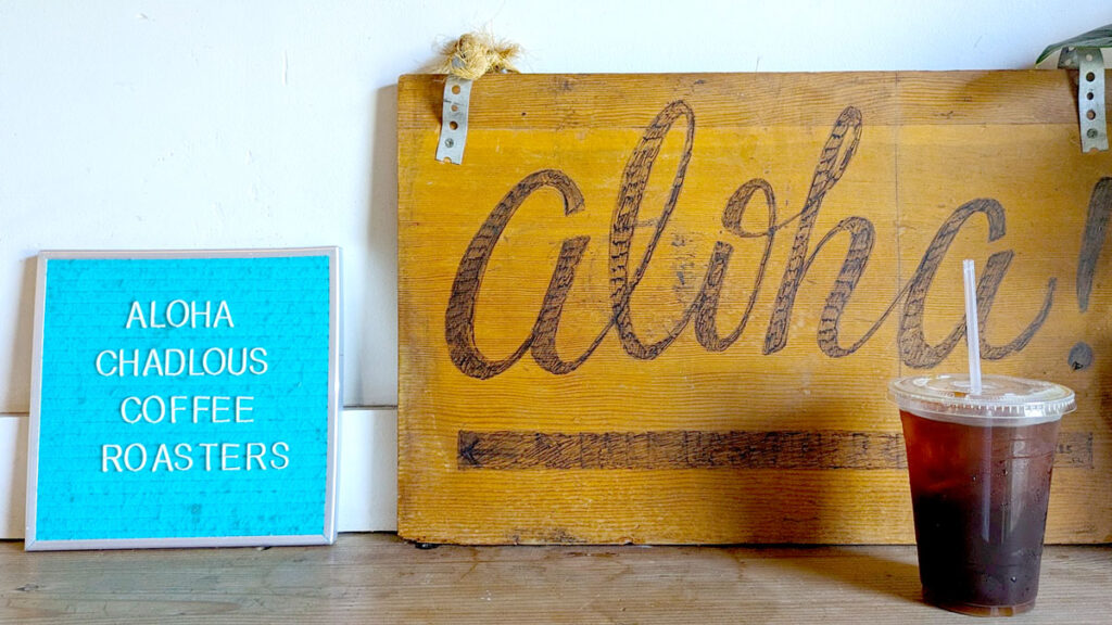 a simple image with three subjects all on a counter top with a white wall in the background. On the left is a bright blue letter board that says: Aloha Chadlous Coffee Roasters. Next to it is a larger wooden board that has Aloha painted on it. And finally there's an iced coffee in a to go cup on the end of the counter.