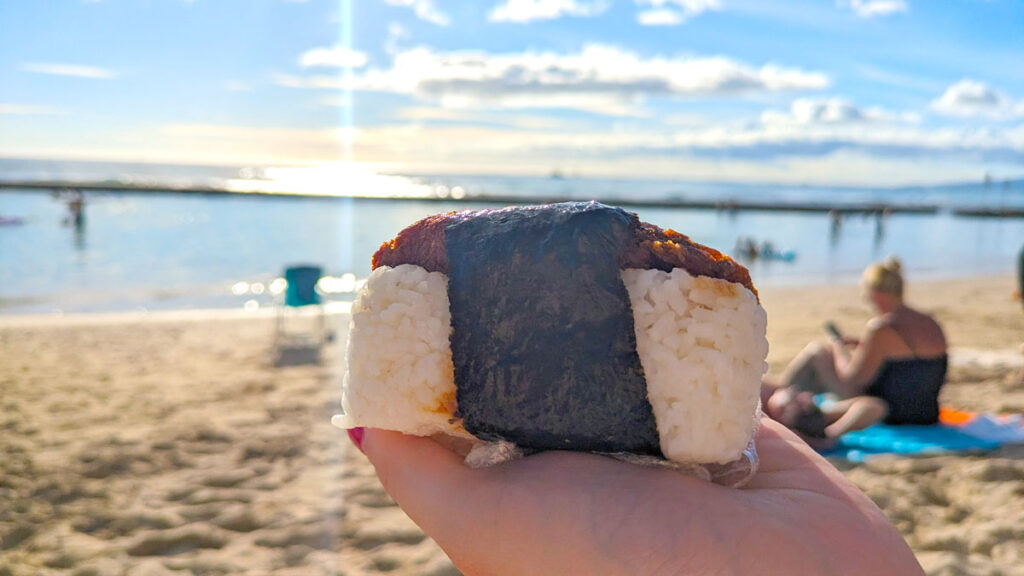 a hand holding up a chicken katsu musubi. A rectangle of densely packed rice with a strip of chicken katsu resting on top. A thin piece of nori wraps around it keeping it all together. The background is the beach at sunset. People are sitting on the sand and are in the water which looks quite walk. The sun is just about to set on the horizon, give the image a wonderful golden hue