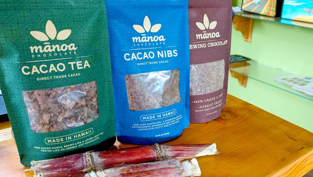 three bags from manoa chocolate on the table. The first bag is green and says cocao tea, you can see through the window of the bag it's brown shells. The middle bag is royal blue and is cocao nibs. Finally, the third bag is a maroon and says brewing chocolate on the bag. 