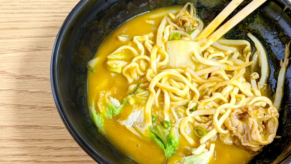 a close up of curry saimin. There's a thick creamy yellow broth with thicker curly saimin noodles pocking out on top and some cabbage. There are wooden chopsticks in the soup