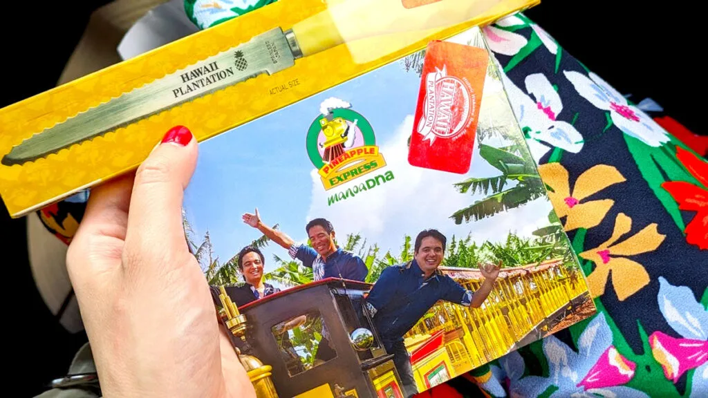 A woman has two items in her lap and she holds them up with one hand. On the top is a serrated knife package. The package shows what the knife looks like inside. It looks more like a mini chainsaw than a knife before it's serrated on both sides. On the knife it says Hawaii Plantation. It has a yellow handle. Below the knife package is a CD called the Pineapple Express by the band Manoa DNA. The CD cover has three men hanging out of a mini yellow train.