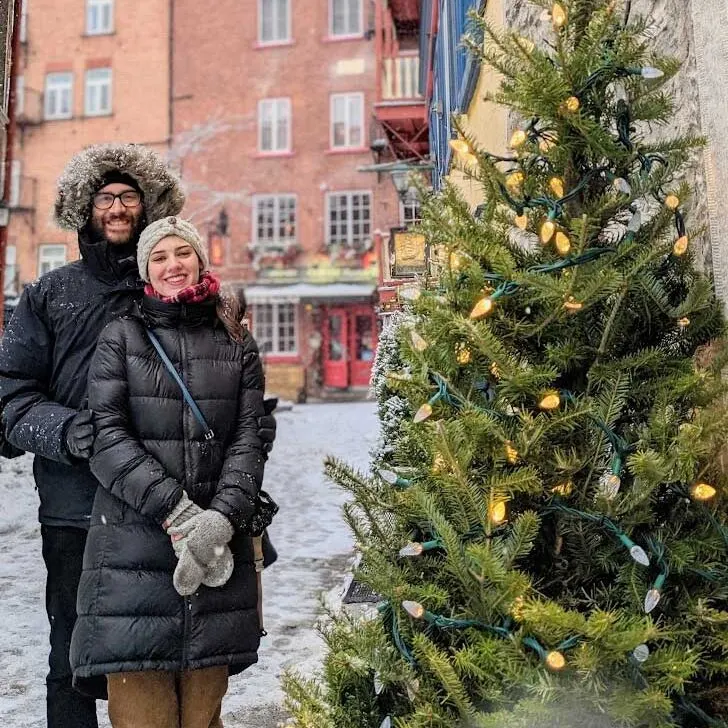 a winter image of quebec city. A woman stands slightly in front of a man, they're both dressed for winter. The mans fur lined hood is over his head for warmth. The woman is wearing a hat and scarf. Both have gloves and winter jackets. They are standing in front of the most iconic view in quebec city: the view of the chateau frontenac from the lower streets in quebec city. There's even a christmas street in front of the couple and a lantern on the side stone wall adding a christmas ambiance to the photo. There's snow on the pavement and it's snowing