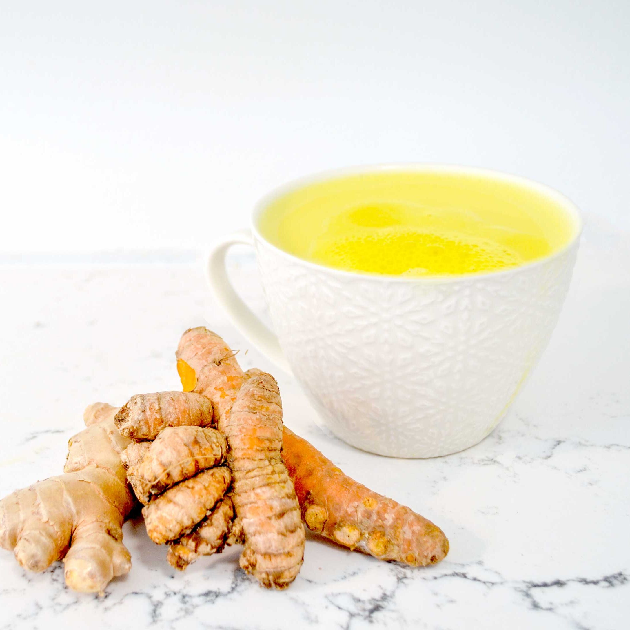 a simple photo of a marble counter top design with a white on white patterned embossed cappuccino mug. There's a bright yellow drink inside, a turmeric tea latte. The are two fresh ungrated stalks of vibrant orange turmeric leaning on each other and the mug and against a similar shaped but lighter in colour root, this is ginger. 