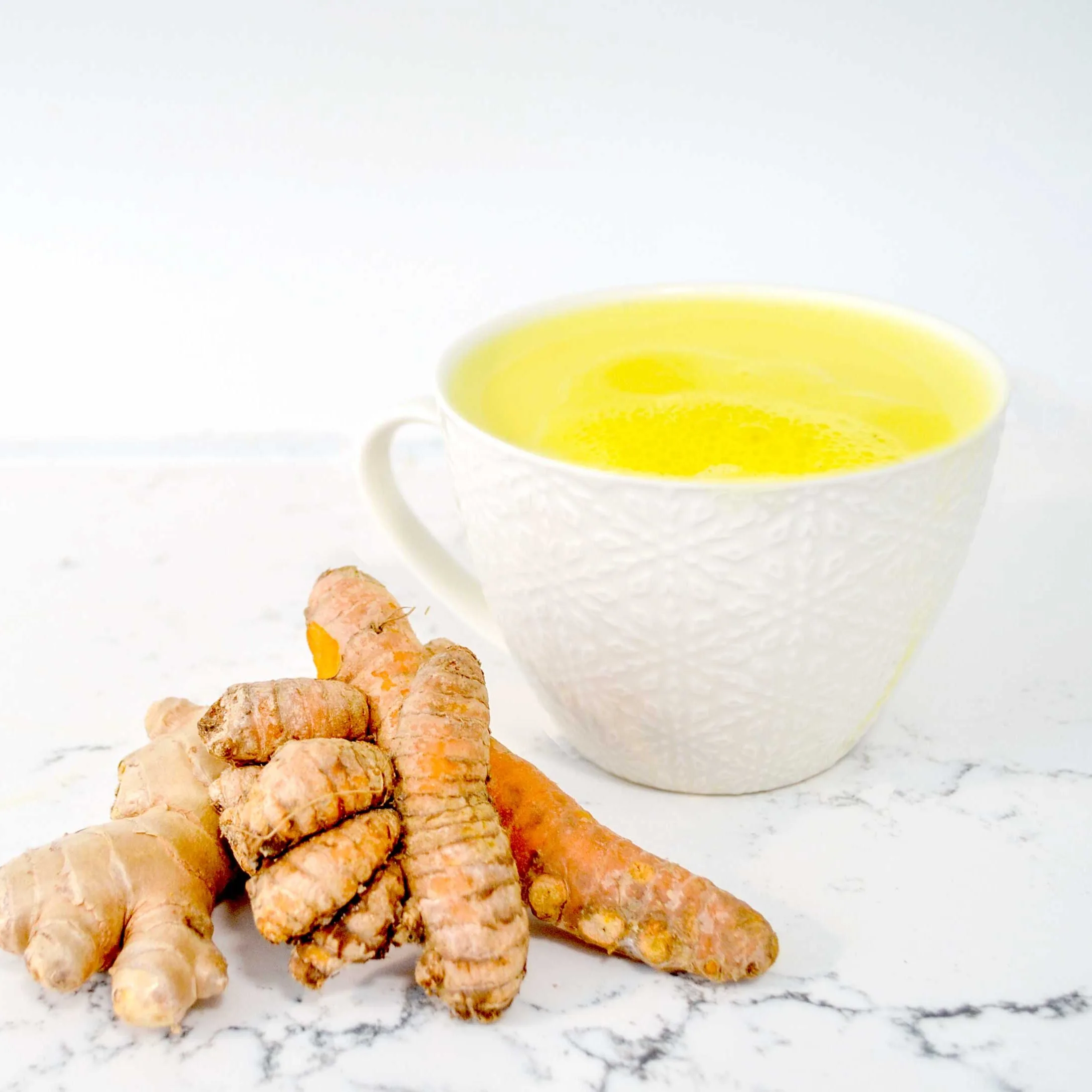 a simple photo of a marble counter top design with a white on white patterned embossed cappuccino mug. There's a bright yellow drink inside, a turmeric tea latte. The are two fresh ungrated stalks of vibrant orange turmeric leaning on each other and the mug and against a similar shaped but lighter in colour root, this is ginger. 