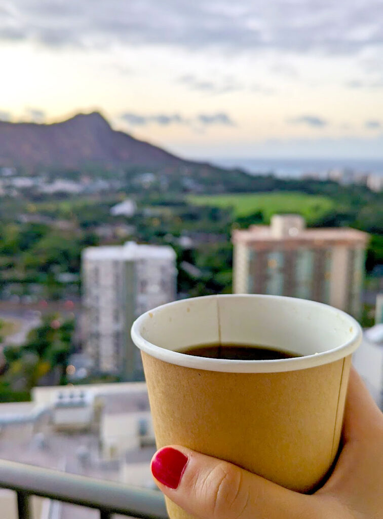 a hand holding a to go cup filled with black coffee in front of building roof tops below and the silhouette of diamond head crater in the distance. There's a yellow purplish hue to the photo and the sky as it's sunrise