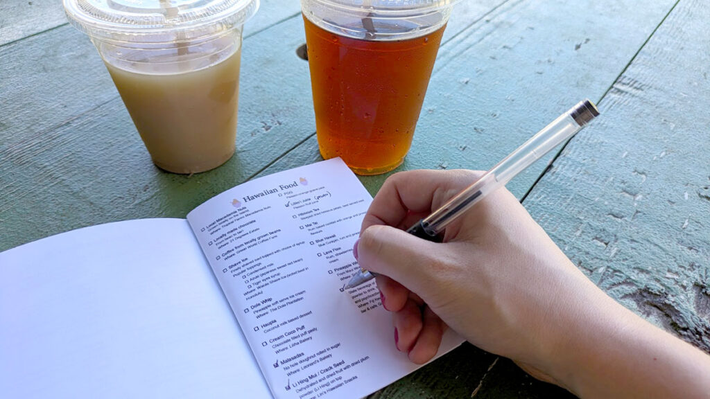 two ice tea drinks on a green picnic table. A booklet is open one the right is a checklist for hawaiian foods and a hand comes into the shot, checking off one of the items on the list