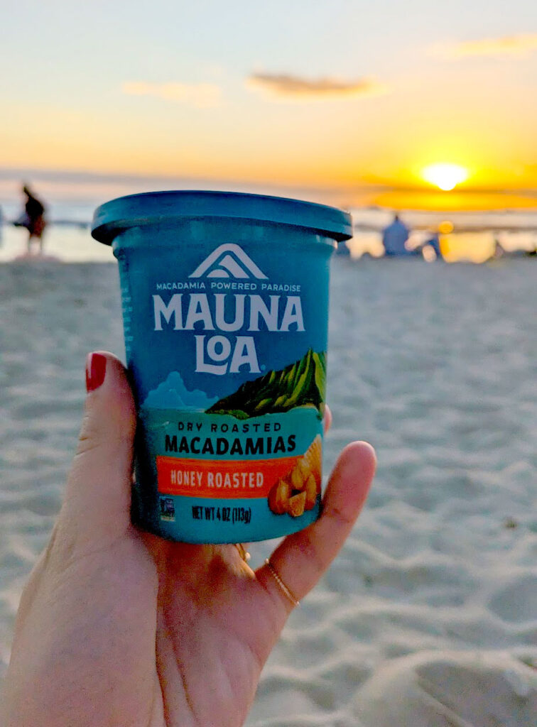 the ultimate hawaiian snack. a tupperware Macadamia nut package held up by a hand in the centre of the photo. The nuts are from Mauna Loa, in a blue package, this is honey roasted flavour. The photo is taken at golden hour on the beach, adding a shadow to the package but an ambiance to the photo. You can see the sand on the bottom, people walking in the middle and finally the ocean and the setting sun before your eyes are lead to the sky.  