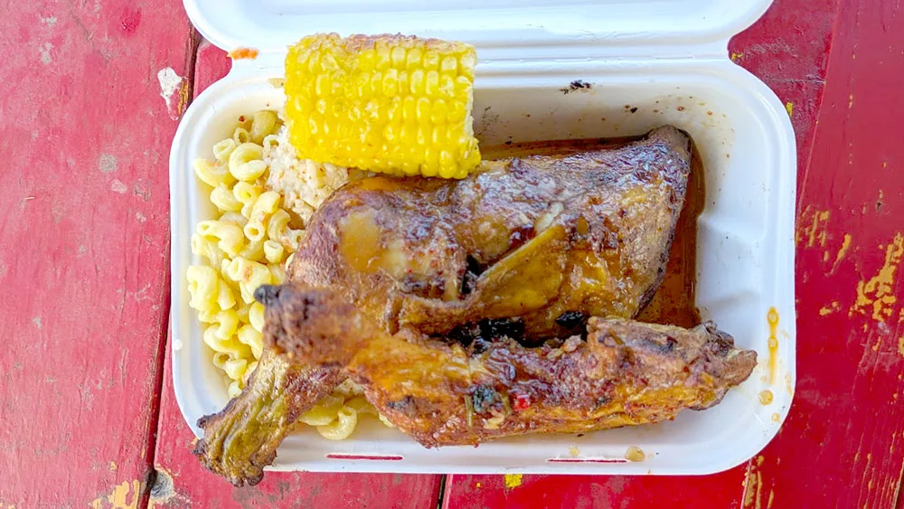 an overhead view of a container with lunch. Huli huli chicken looks well roasted in a sauce making the chicken a deep brown and you can see speckles of spices. It sits on top of a traditional mac salad, and there's a quarter of a piece of corn on the cobb. The container is on a painted red picnic table that has some chips in the paint. 