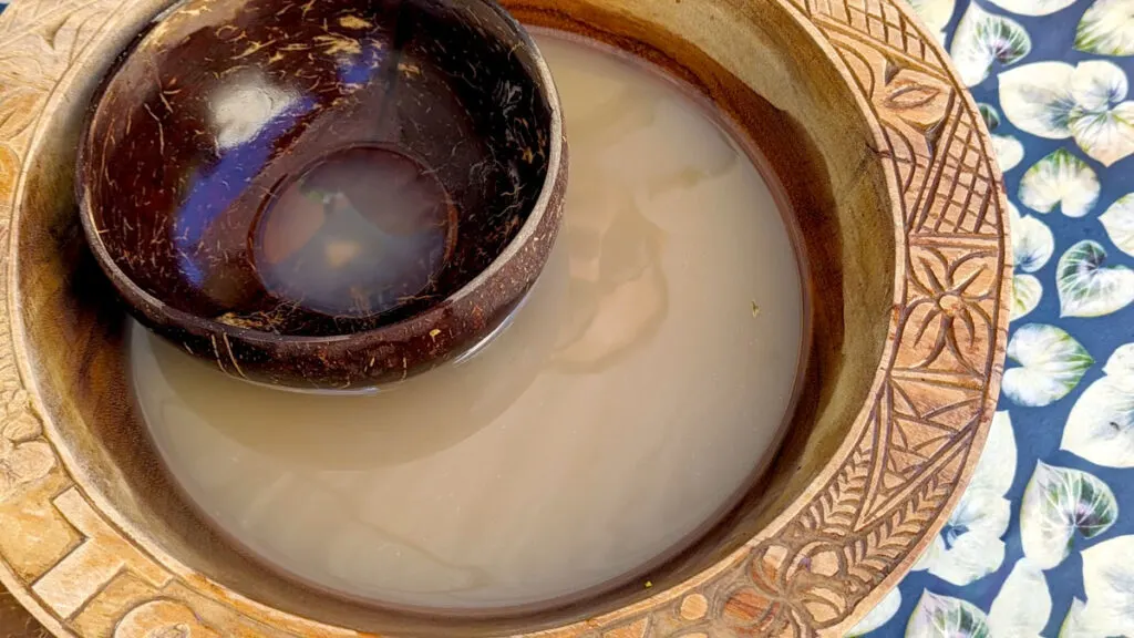 a traditional large kava bowl sits on a blue and white patterned table cloth. The edges of the large bowl have etchings on the rim. Inside it's filled with a light brown liquid: kava. And inside that is a small coconut like shaped bowl for serving.