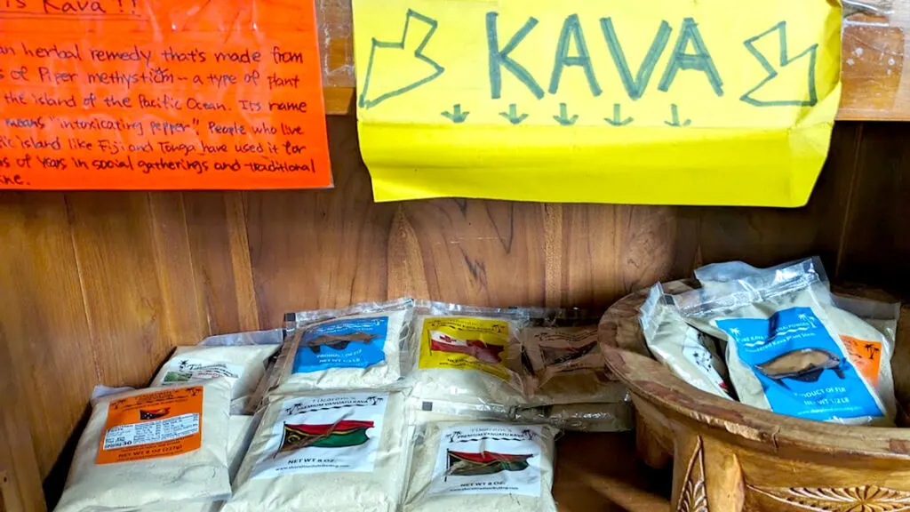 A wooden shelf that has posters taped to the top shelf made from coloured paper and written on with market. The firs sheet is orange and describes what kava is, but you can't really see what's written. The second is bright yellow, and says KAVA in giant letter with large arrows pointing below. On the bottom shelf are a few bags filled with a brown powder. 