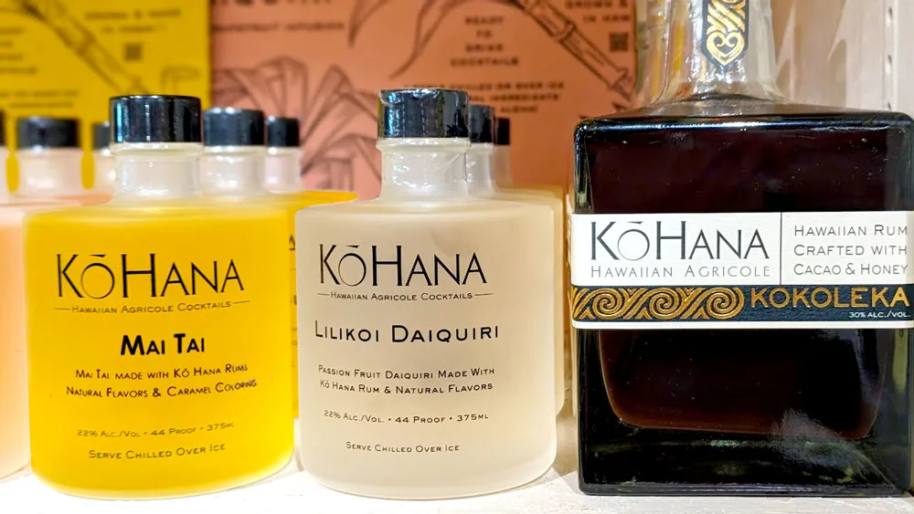 Three bottles on a shelf from the Ko'hana distilleries. The first bottle is frosted glass but you can still see the amber brew inside. It's a mai tai mix. The second is slightly more white, and it's a liliko'i mix .The last bottle is different, it's square and it's simply Hawaiian rum.