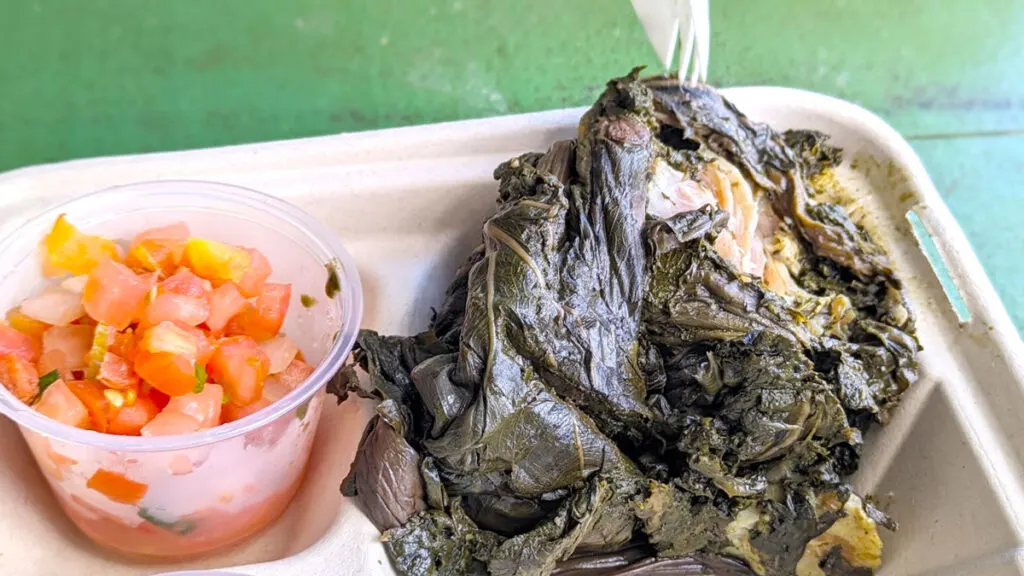 a closeup of traditional hawaiian food: lomi-salmon and pork laulau. 
The lomi salmon is in a small plastic cup, it looks like finely chopped tomatoes. Next to it is the laulau, not as good looking in presentation. It's wet taro leaves that are wrapped around a meat you can see in a small corner where the taro leaves have been ripped open by a fork. 