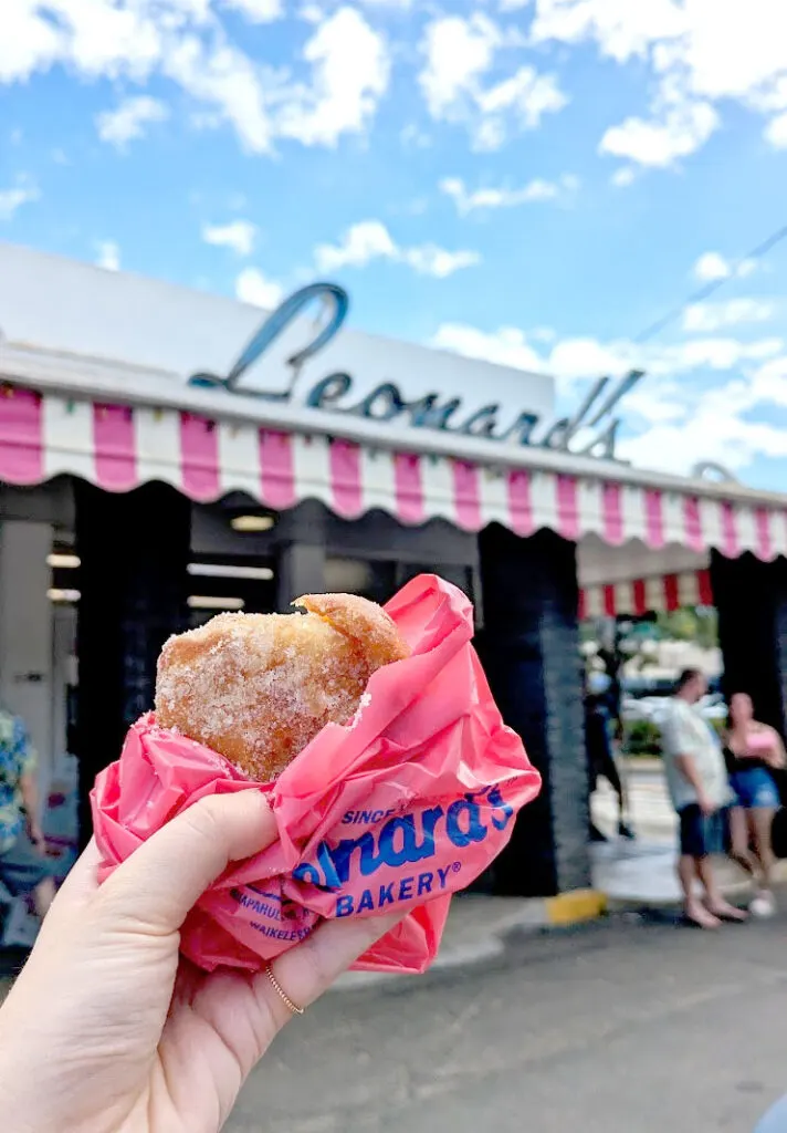 a hand holding up a bright pink paper bag showing a fried sugar covered doughnut inside. In the background is a pink and white awning with vintage sign for Leonards on top. It's a sunny day but there are a few clouds in the sky that add movement and brings you eye downward towards the shop.