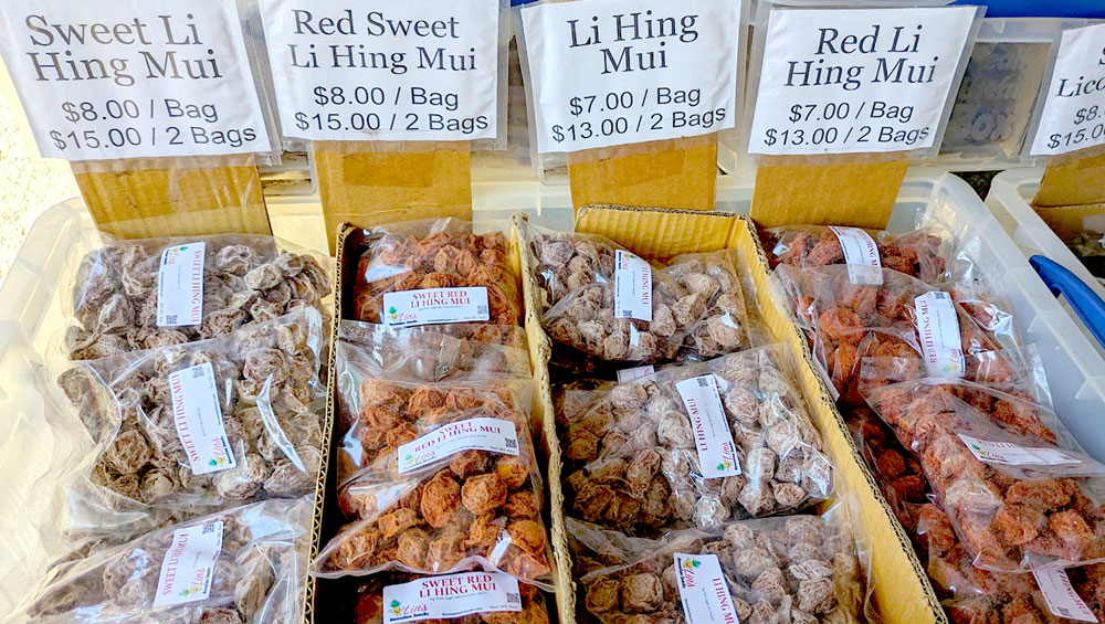 a box filled with bags of crack see at a hawaiian market. The labels on top of the box are printed at home. There are four rows of different types of dried li hing mui fruit. They're dark and bright reds
