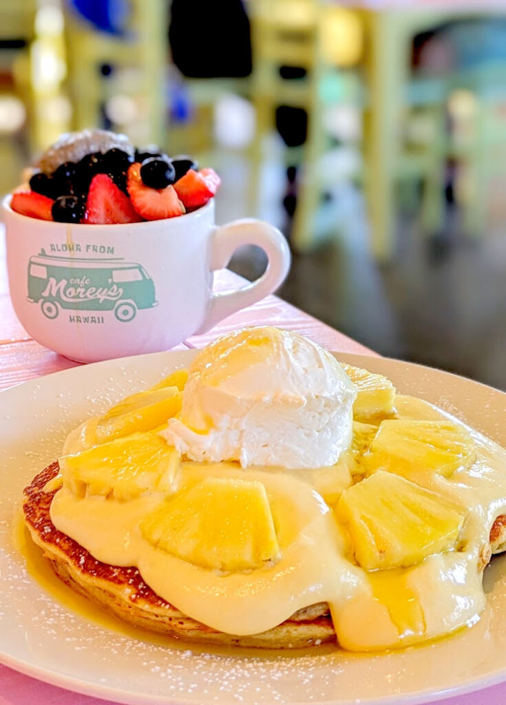 A very bright image of fun colours and delicious Hawaiian desserts on a pink table. A large plate is in the foreground, taken over by a huge pancake that you can barely see under a yellow cream, pineapple slices, and a dollop of whipped cream to top it all off. But that's not all for breakfast. There's a large "friends" style mug behind the plate with a light teal Morey's Cafe logo of a van. The cup is filled with bright red and blue berries and chia seed pudding. 